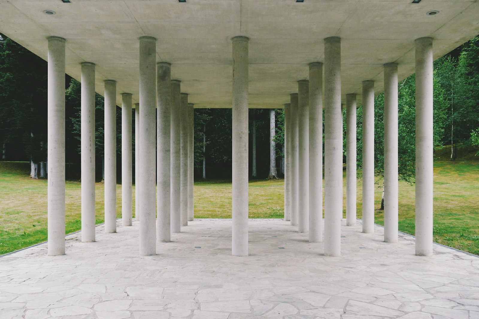 Rows of plain concrete columns under a flat roof on stone paving, with a lawn and trees behind
