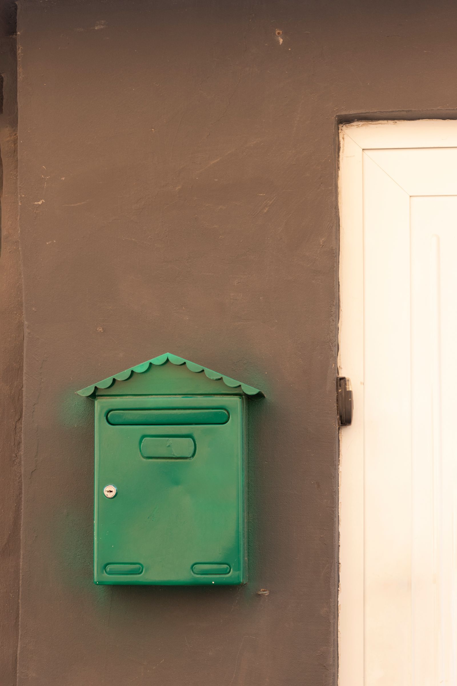 A green metal mailbox mounted on a brown plaster wall beside a white door