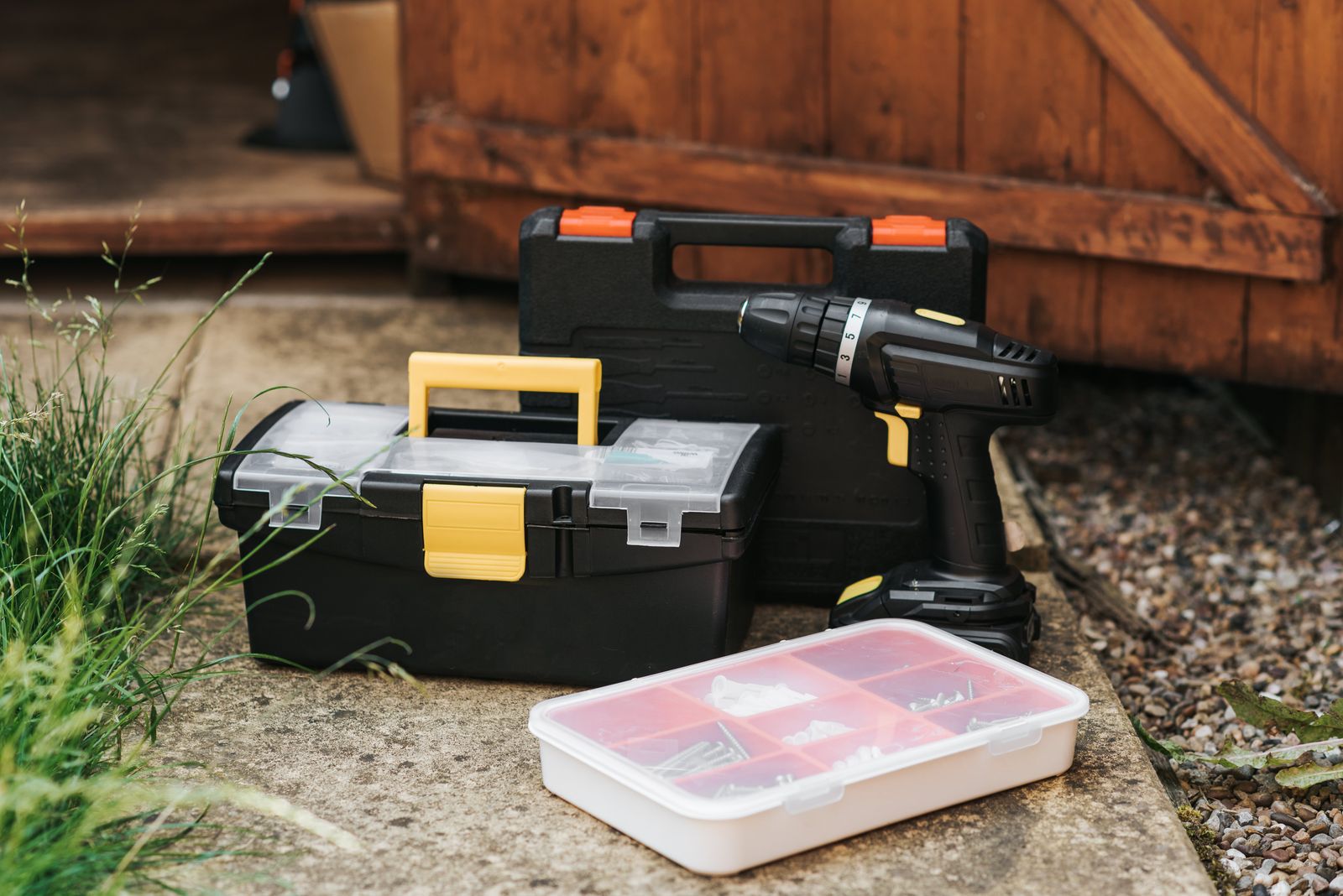 Two black toolboxes, a cordless drill, and a parts box set down on a paved path beside a wooden gate